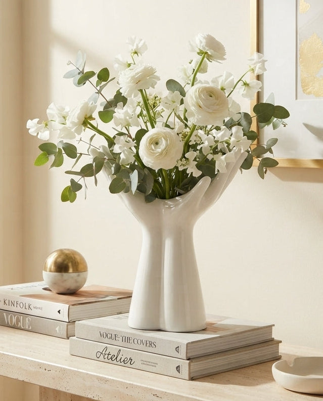 A white ceramic vase shaped like a hand holding flowers, filled with white roses and green eucalyptus, styled on a light wood console table with books and a small gold sphere in a bright, warm interior setting.